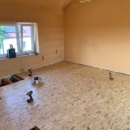 Montaż podłogi z płyty OSB w pokoju z oknem, widoczne narzędzia: wkrętarki, poziomica, puszka z wkrętami, fragment instalacji elektrycznej przy ścianie