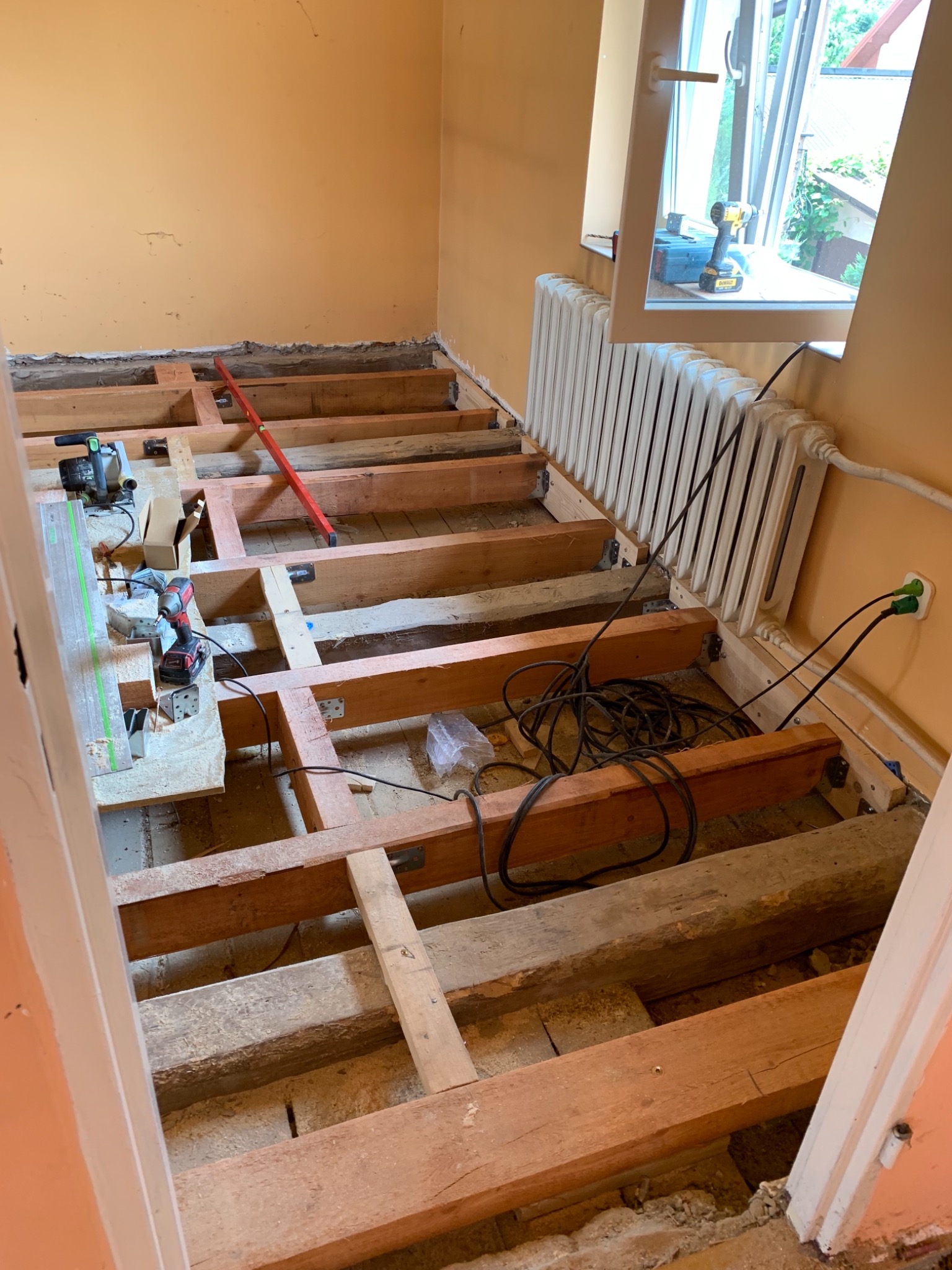 Przygotowanie podłoża pod nową podłogę: drewniana konstrukcja legarów w pokoju z grzejnikiem i narzędziami, widok z progu drzwi.