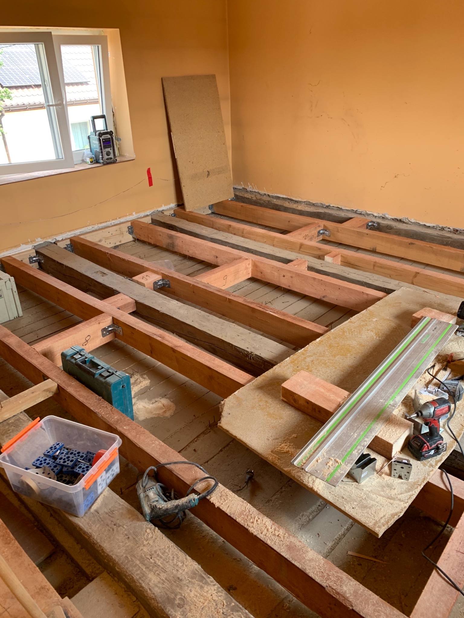 Wnętrze w trakcie remontu podłogi: drewniane legary ułożone na starym parkiecie, narzędzia pracy (wiertarka, szyna prowadząca, skrzynka z łącznikami) rozłożone na podłodze, ściany w kolorze...