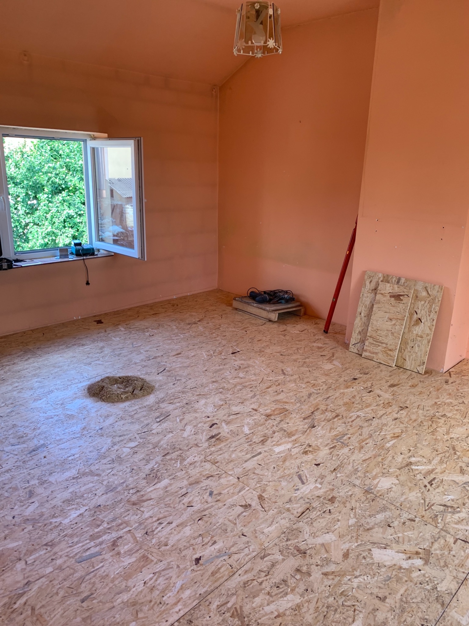 Pokój w trakcie remontu z wyłożoną płytą OSB na podłodze, ścianami w kolorze brzoskwiniowym, narzędziami na palecie i otwartym oknem z widokiem na zieleń.