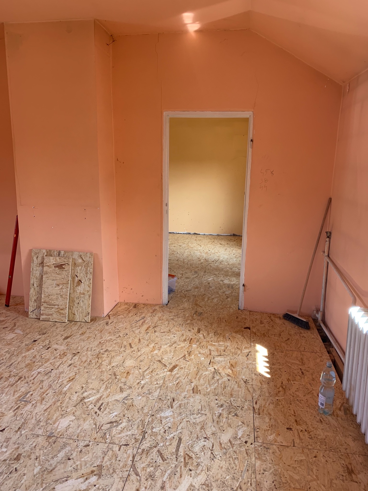 Przygotowanie podłoża z płyt OSB pod dalsze prace wykończeniowe w pokoju w Tarnobrzegu, widoczne płyty na podłodze, fragment ściany pomalowanej na łososiowy kolor oraz butelka wody.