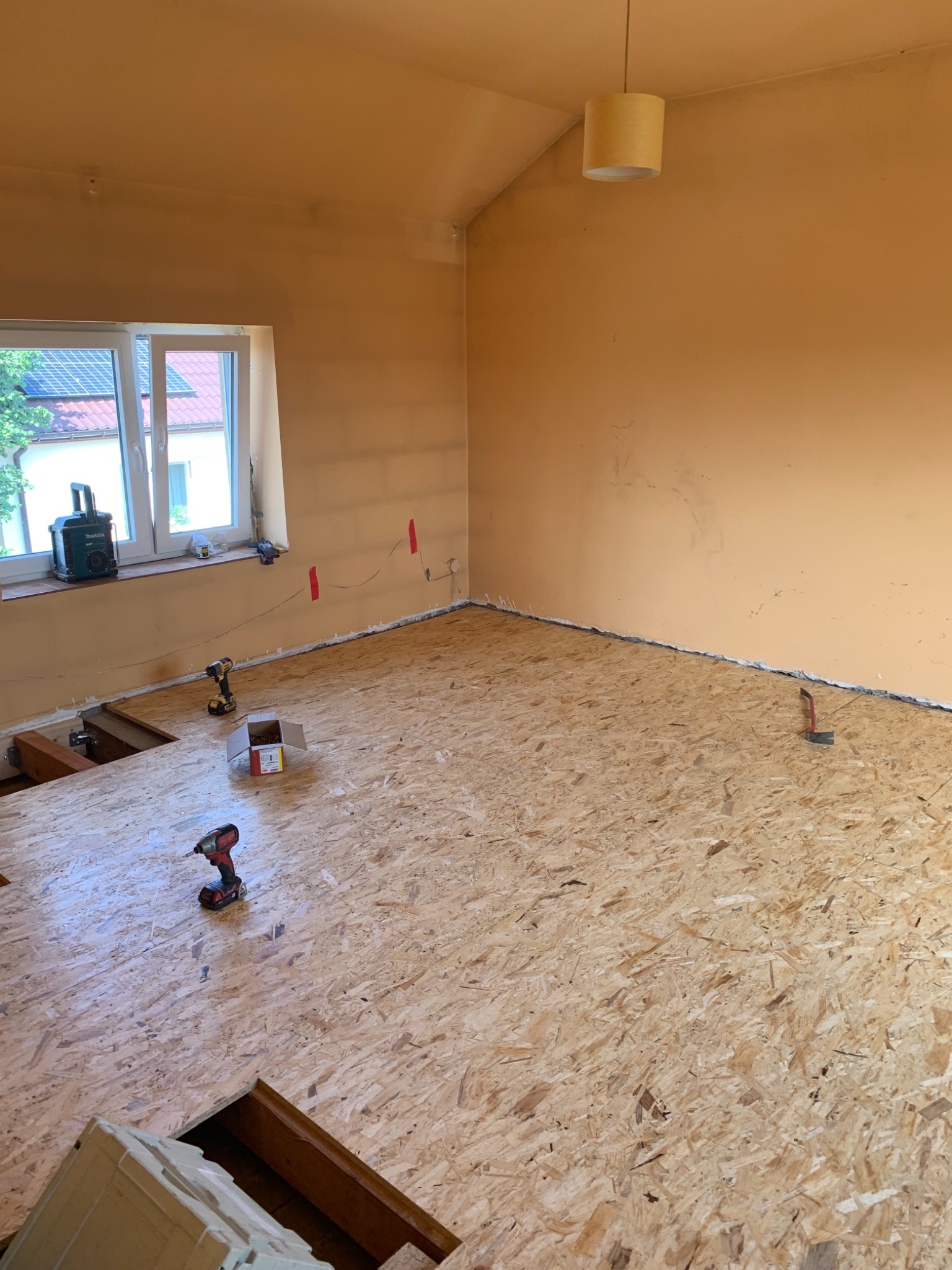 Montaż podłogi z płyty OSB w pokoju z oknem, widoczne narzędzia: wkrętarki, poziomica, puszka z wkrętami, fragment instalacji elektrycznej przy ścianie