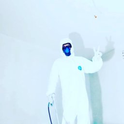 Tomdeco - Pracownik w kombinezonie ochronnym i masce, z pistoletem natryskowym, pozuje z dwoma palcami w górze w świeżo pomalowanym, jasnym pomieszczeniu.