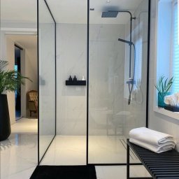 Tomdeco - Nowoczesna łazienka z kabiną prysznicową typu walk-in z czarnymi profilami, marmurowe płytki na ścianach, czarna półka na kosmetyki, czarny dywanik i czarny stolik z ręcznikami.