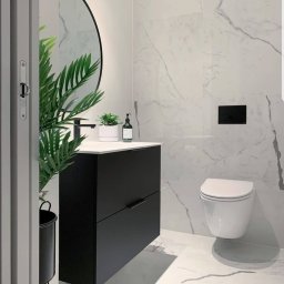 Tomdeco - Nowoczesna łazienka z czarną szafką pod umywalką, okrągłym lustrem z podświetleniem, białymi płytkami imitującymi marmur na ścianach i podłodze oraz wiszącą toaletą.