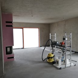 Tomdeco - Wnętrze w trakcie wykańczania: surowe ściany i sufit po gładziowaniu, widoczny kominek obudowany płytami, żółty odkurzacz przemysłowy i stół roboczy na rusztowaniu.