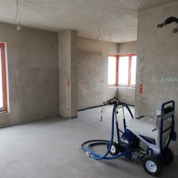 Tomdeco - Wnętrze w trakcie prac wykończeniowych: aplikacja gładzi maszynowej, widoczny agregat malarski Texspray T-Max na tle surowych ścian i wnęk okiennych z pomarańczowymi ramami. Na ścianie widnieją...
