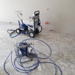 Tomdeco - Dwa agregaty malarskie Graco, jeden na wózku z dużymi kołami, drugi mniejszy z długim wężem, stoją w pustym, surowym pomieszczeniu z widocznymi puszkami instalacyjnymi.