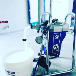 Tomdeco - Profesjonalny agregat malarski Graco GX 21 z białym gruntem Flügger Hall Primer White w wiadrze, przygotowany do malowania ścian w pomieszczeniu zabezpieczonym taśmą malarską.