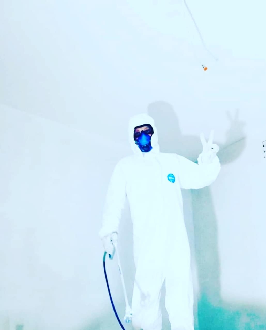 Pracownik w kombinezonie ochronnym i masce, z pistoletem natryskowym, pozuje z dwoma palcami w górze w świeżo pomalowanym, jasnym pomieszczeniu.