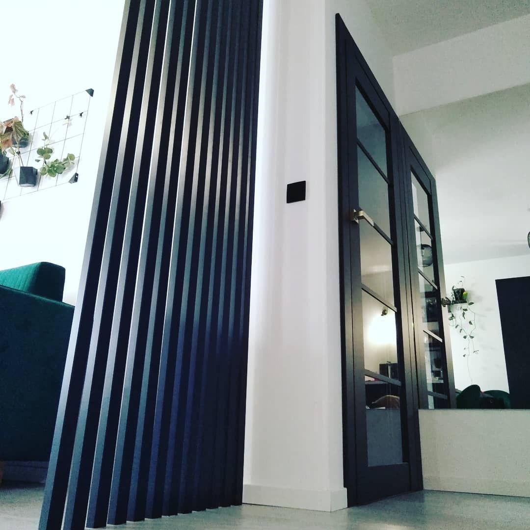 Nowoczesny salon z czarnymi, pionowymi lamelami jako element dekoracyjny, biała ściana z czarnym włącznikiem światła, przeszklone, ciemne drzwi dwuskrzydłowe.