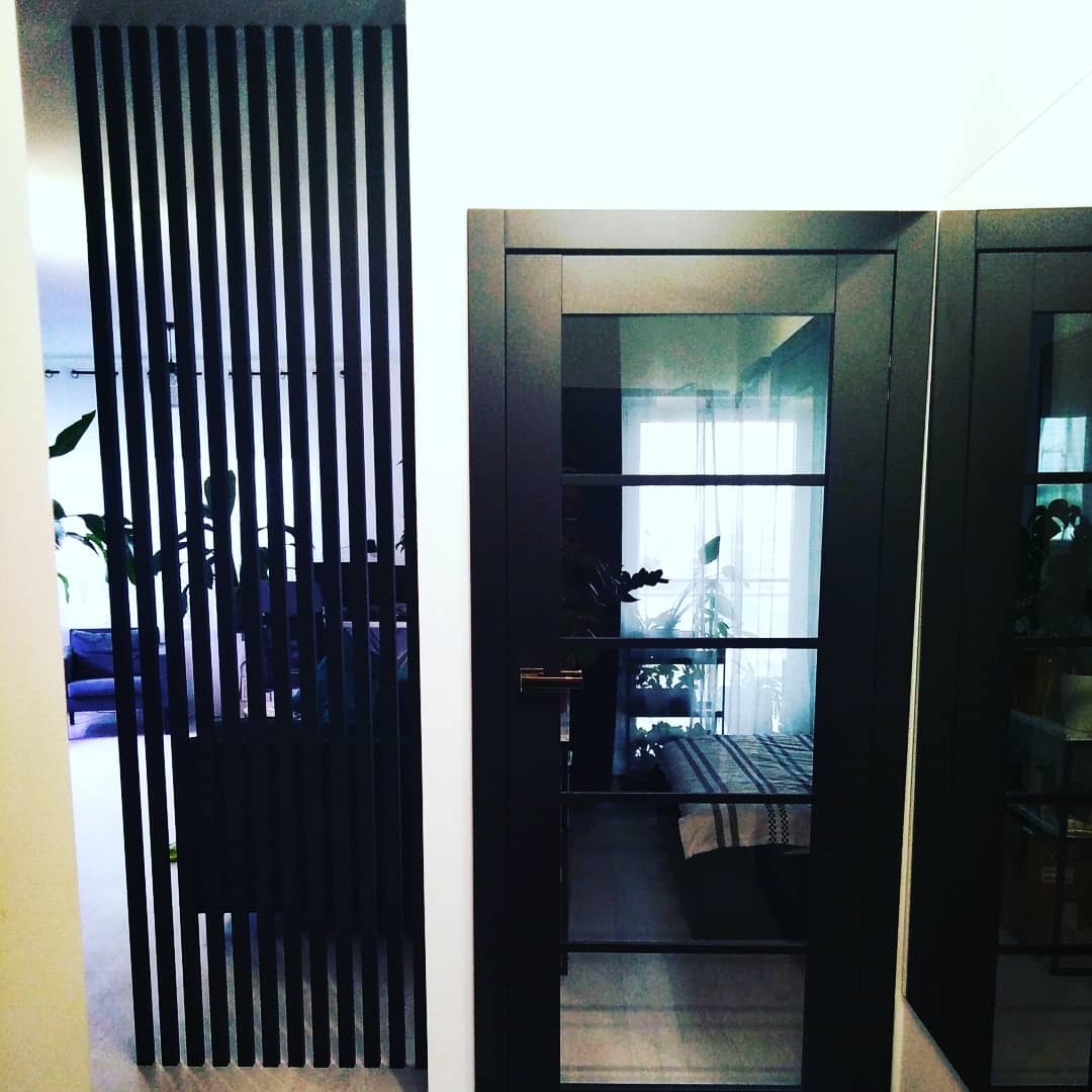 Nowoczesna aranżacja wnętrza z czarną, ażurową ścianką działową i drzwiami z przeszkleniami, widok na salon z sofą i roślinami.