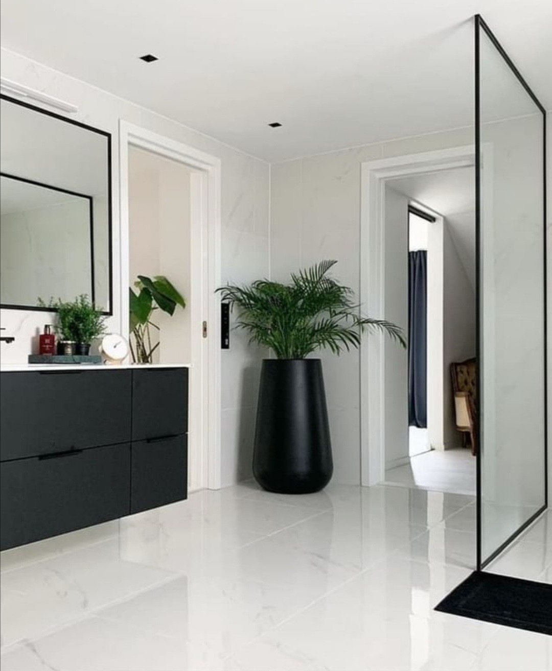 Nowoczesna łazienka z białymi marmurowymi płytkami, czarną szafką pod umywalką, lustrem w czarnej ramie, dużą rośliną w czarnej donicy i szklaną kabiną prysznicową typu walk-in z czarnymi...