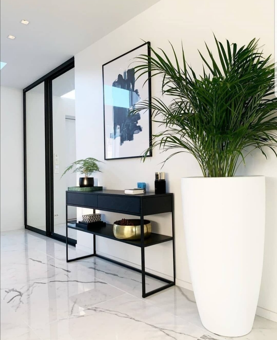 Nowoczesny przedpokój z czarną konsolą, abstrakcyjnym obrazem i dużą rośliną w białej donicy, na podłodze biały marmur z szarymi żyłami.