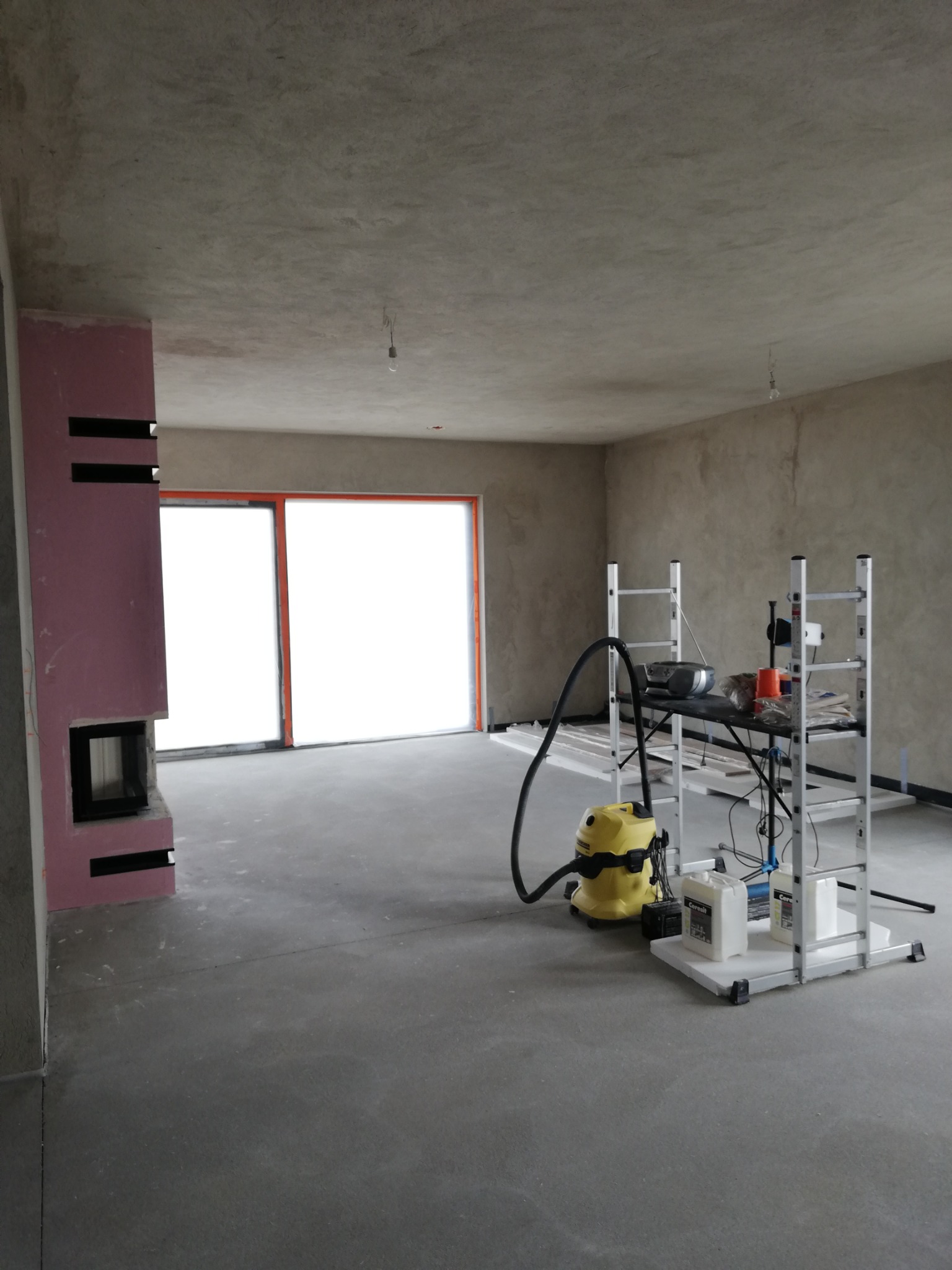 Wnętrze w trakcie wykańczania: surowe ściany i sufit po gładziowaniu, widoczny kominek obudowany płytami, żółty odkurzacz przemysłowy i stół roboczy na rusztowaniu.