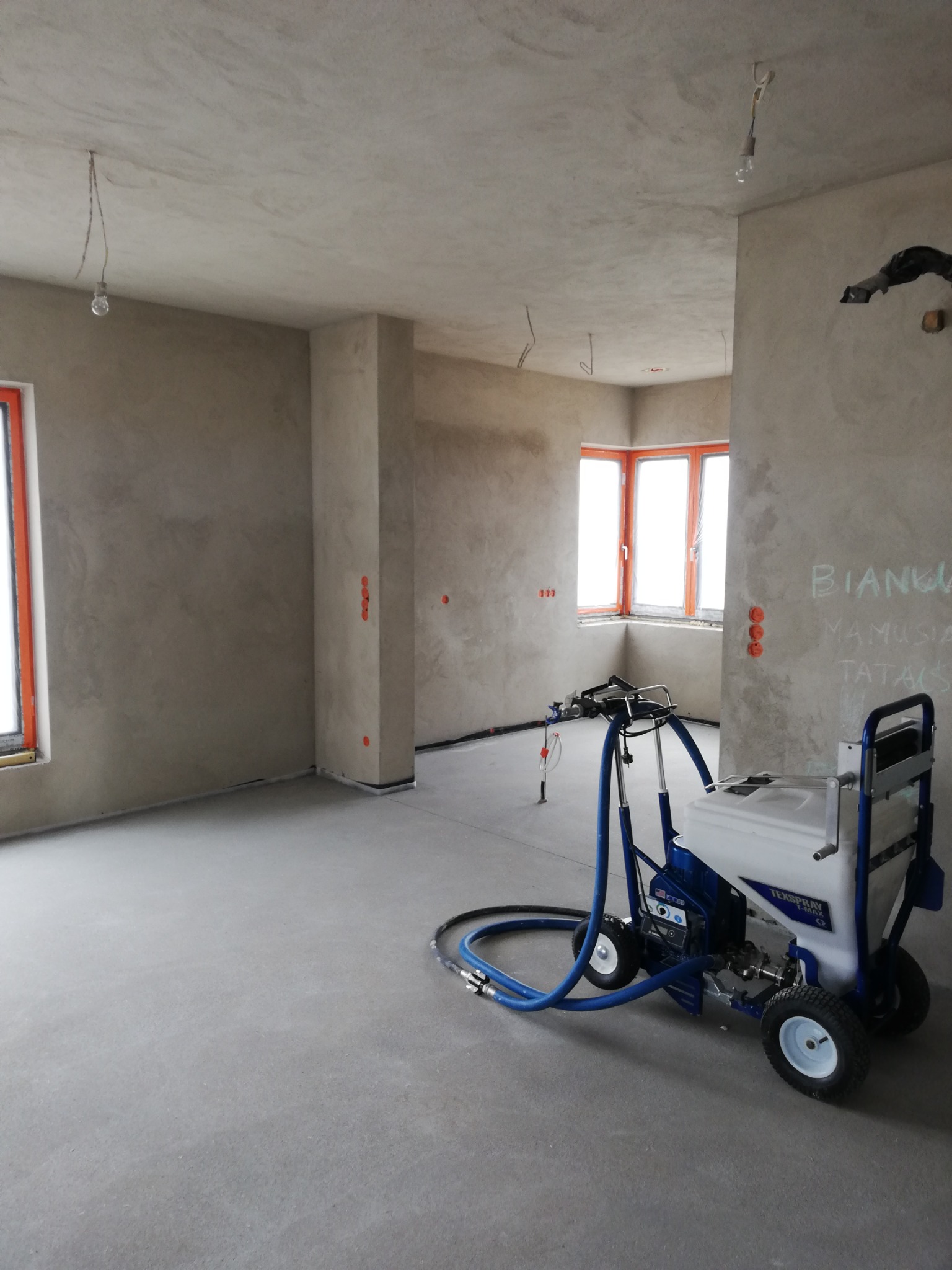 Wnętrze w trakcie prac wykończeniowych: aplikacja gładzi maszynowej, widoczny agregat malarski Texspray T-Max na tle surowych ścian i wnęk okiennych z pomarańczowymi ramami. Na ścianie widnieją...