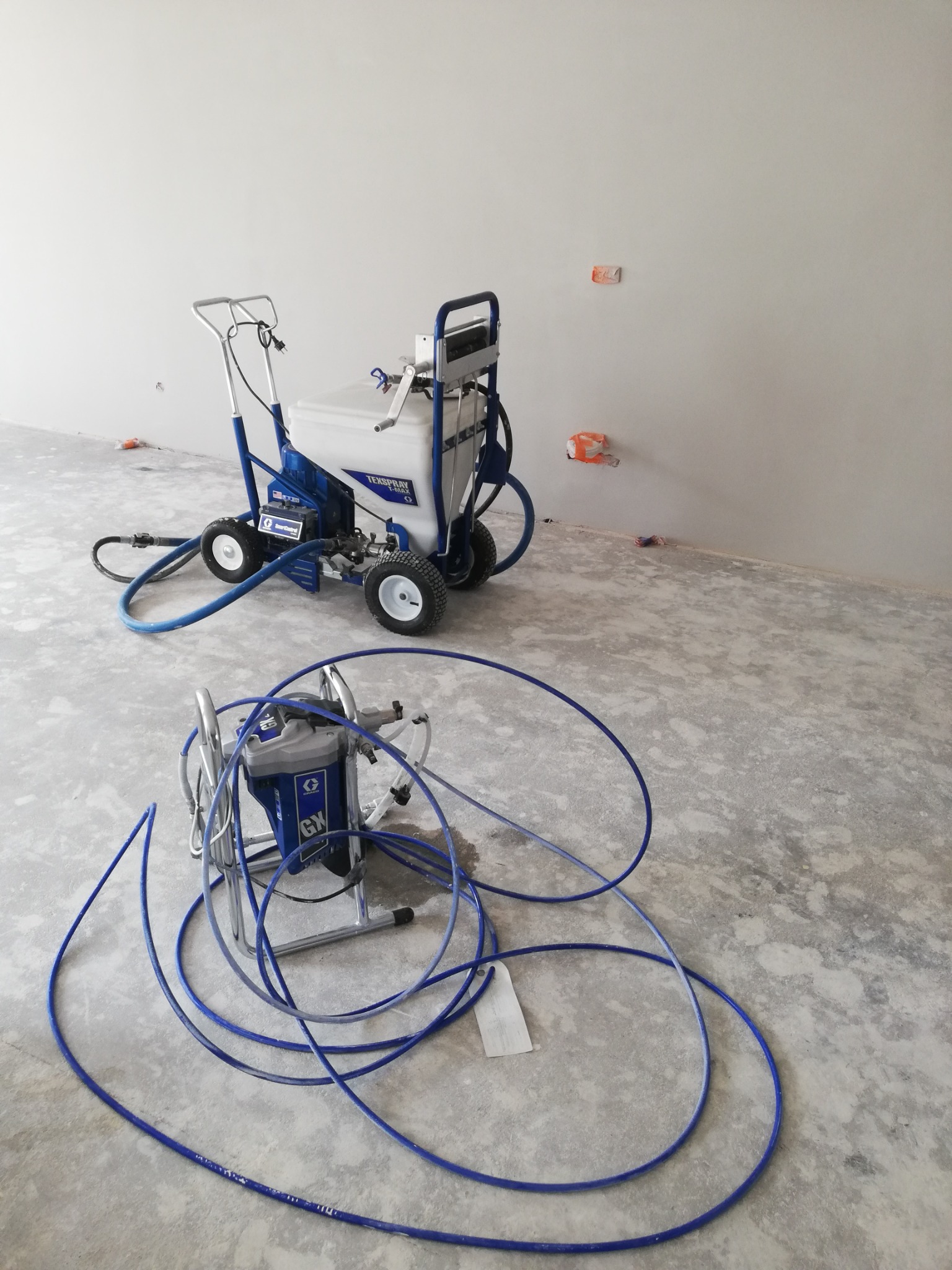 Dwa agregaty malarskie Graco, jeden na wózku z dużymi kołami, drugi mniejszy z długim wężem, stoją w pustym, surowym pomieszczeniu z widocznymi puszkami instalacyjnymi.