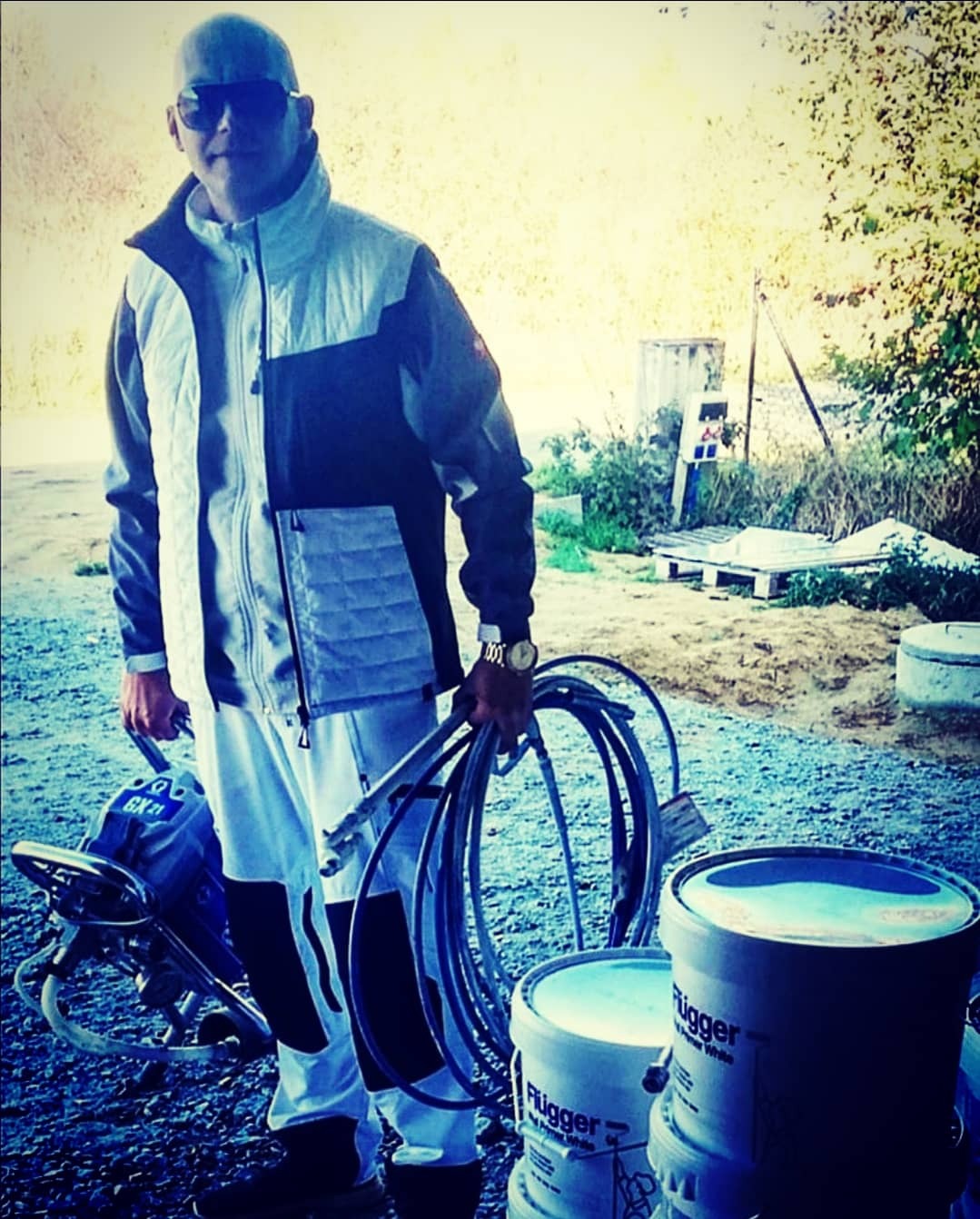 Mężczyzna w okularach przeciwsłonecznych trzymający wąż do agregatu malarskiego, obok stoją wiadra z farbą Flugger White