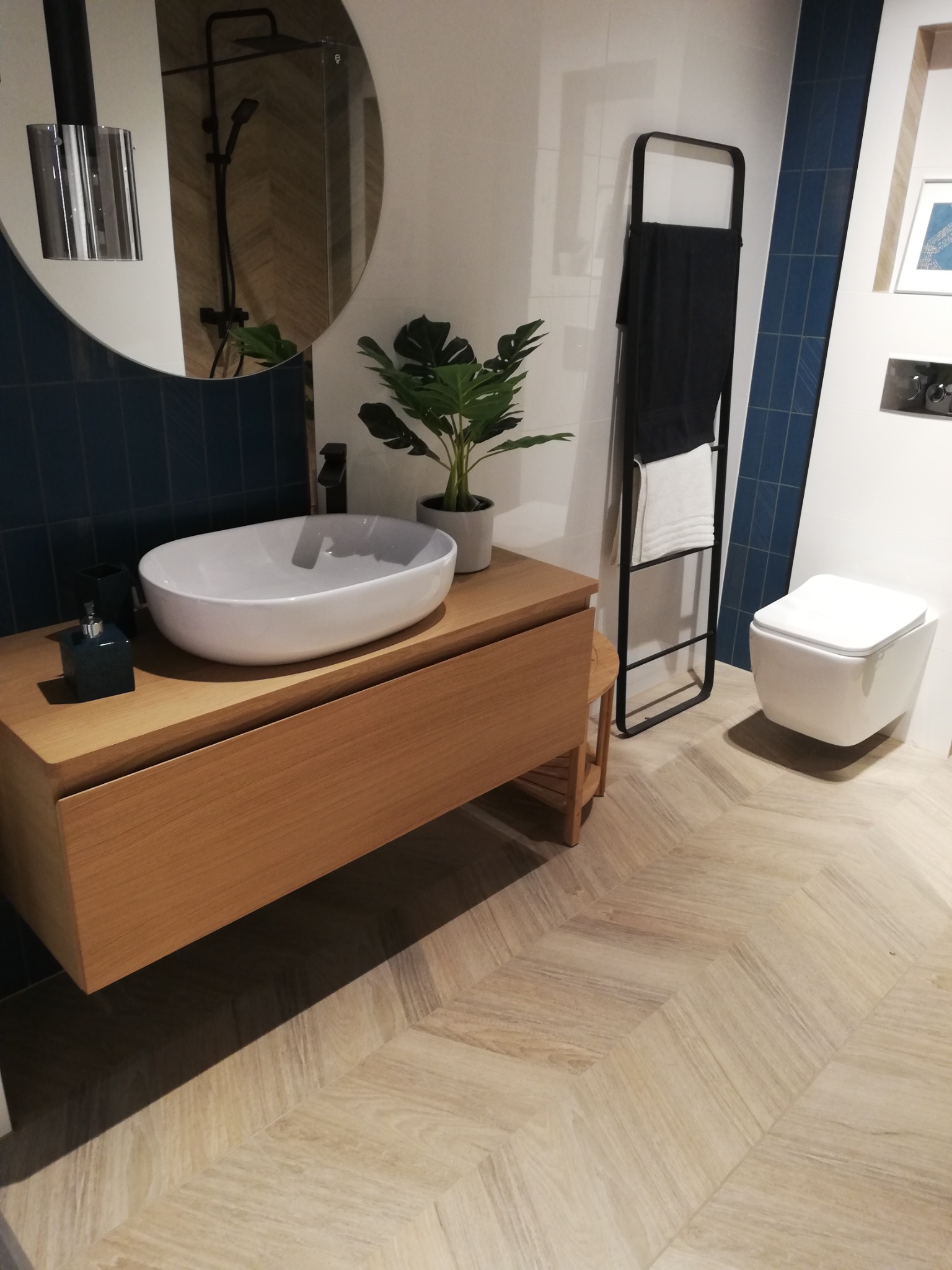 Nowoczesna łazienka z drewnianą szafką pod umywalką, owalną białą umywalką nablatową, czarną armaturą, lustrem w okrągłej ramie, niebieskimi płytkami na ścianie i podłogą ułożoną w jodełkę.