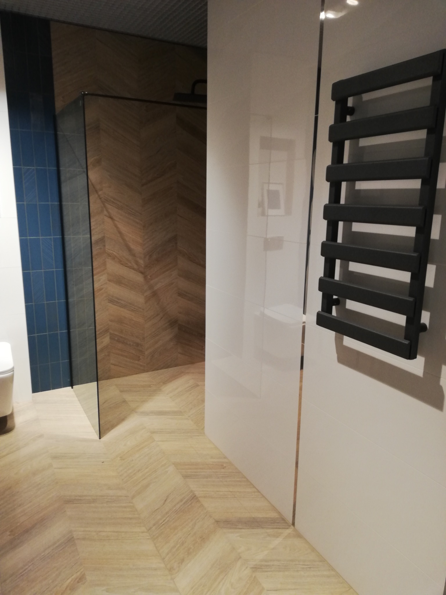 Nowoczesna łazienka z drewnopodobną podłogą ułożoną w jodełkę, kabiną prysznicową ze szklaną ścianką i czarnym grzejnikiem drabinkowym na białej ścianie.