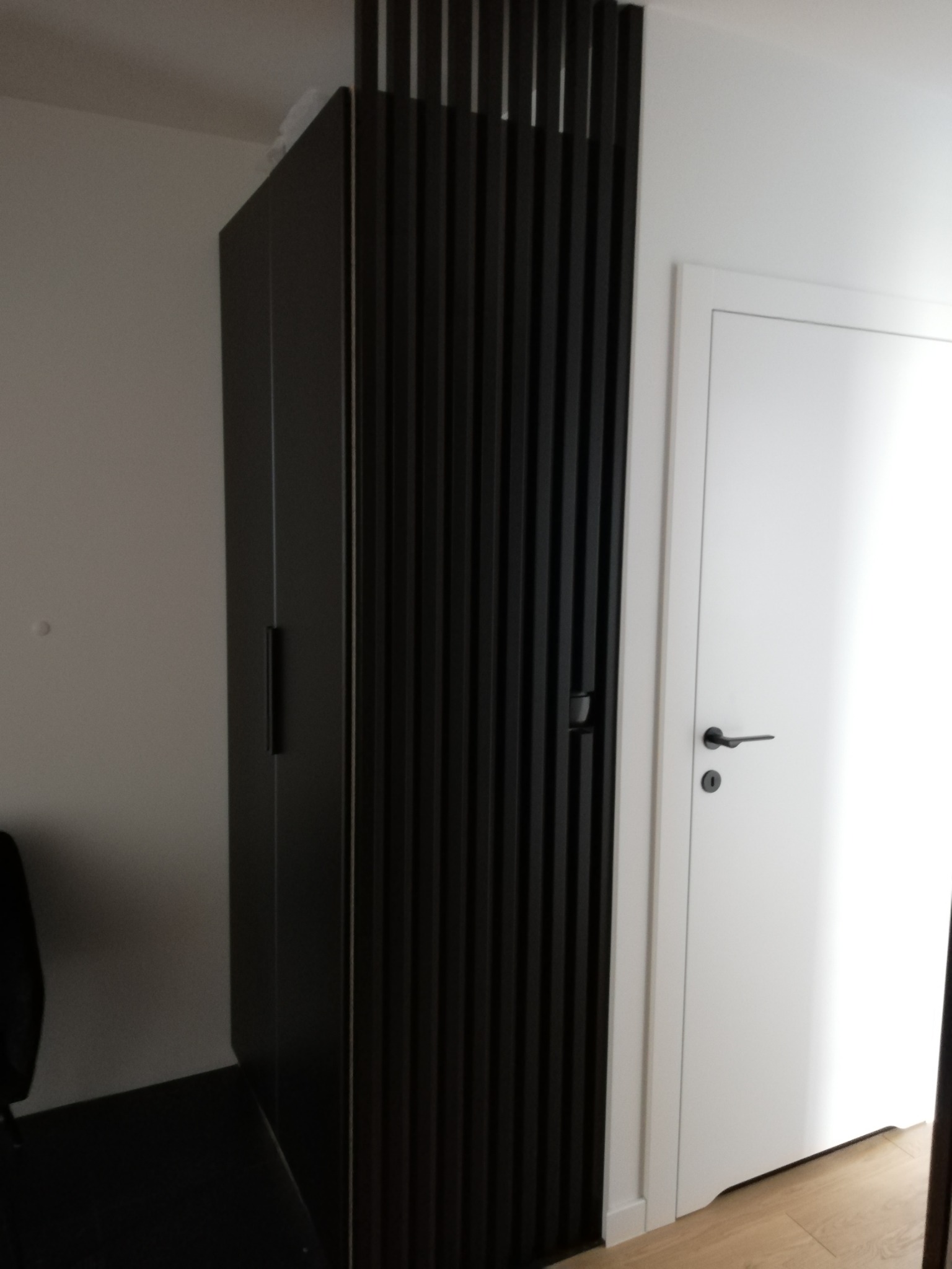 Ciemna zabudowa meblowa z lamelami pionowymi, przylegająca do białej ściany i drzwi w minimalistycznym wnętrzu.