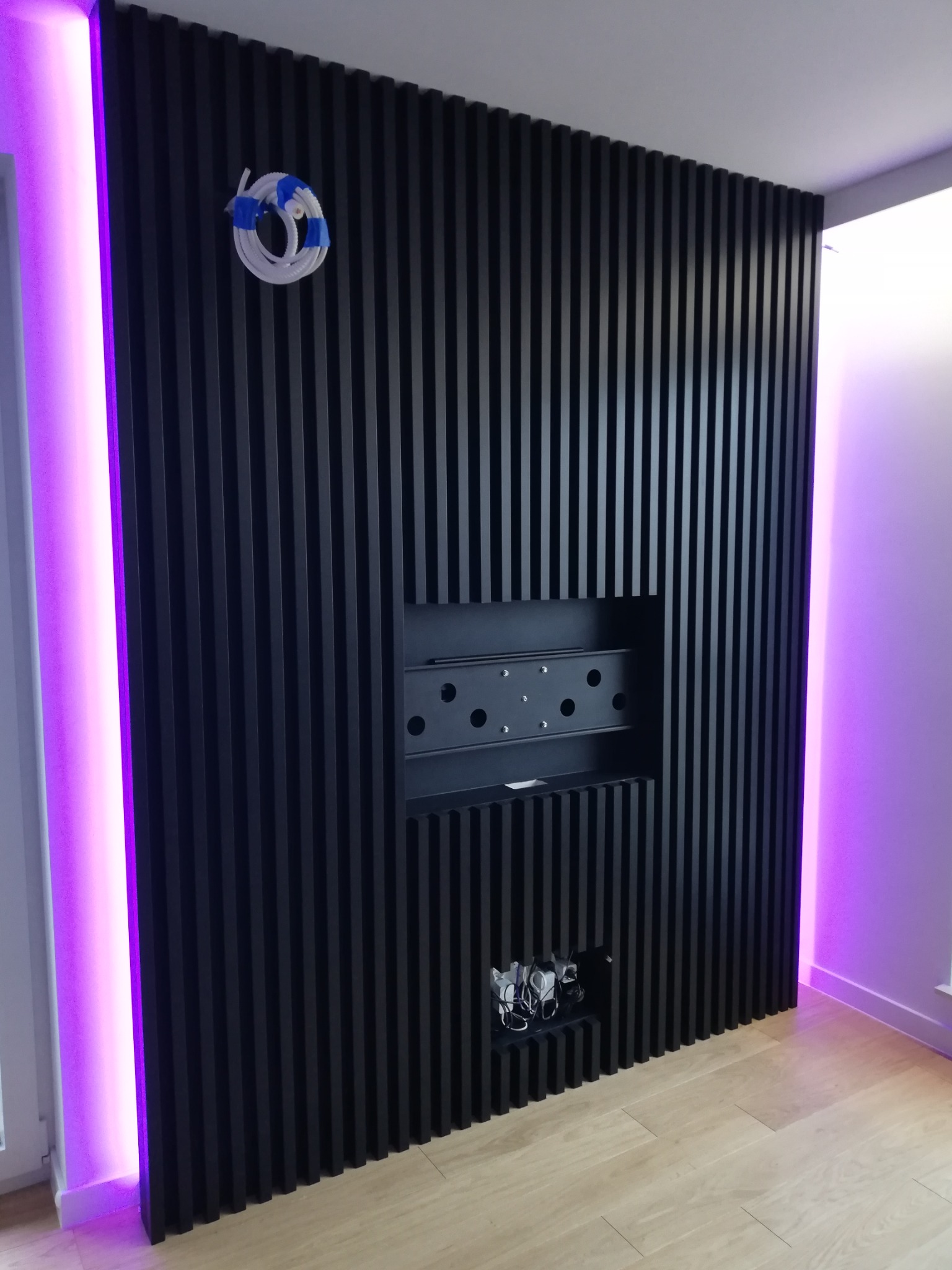Czarny panel ścienny z pionowych listew z wnękami na TV i kable, podświetlony fioletowym światłem LED po lewej stronie.