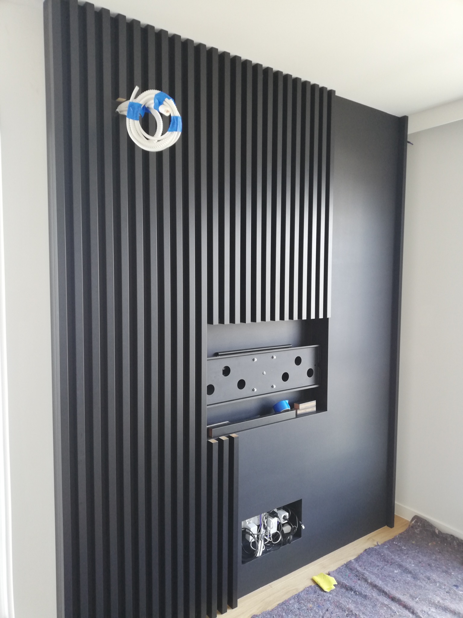 Częściowo ukończona ściana z czarnych lameli z przygotowanym miejscem na telewizor i ukrytymi gniazdkami elektrycznymi, widoczne przewody i zabezpieczona podłoga.