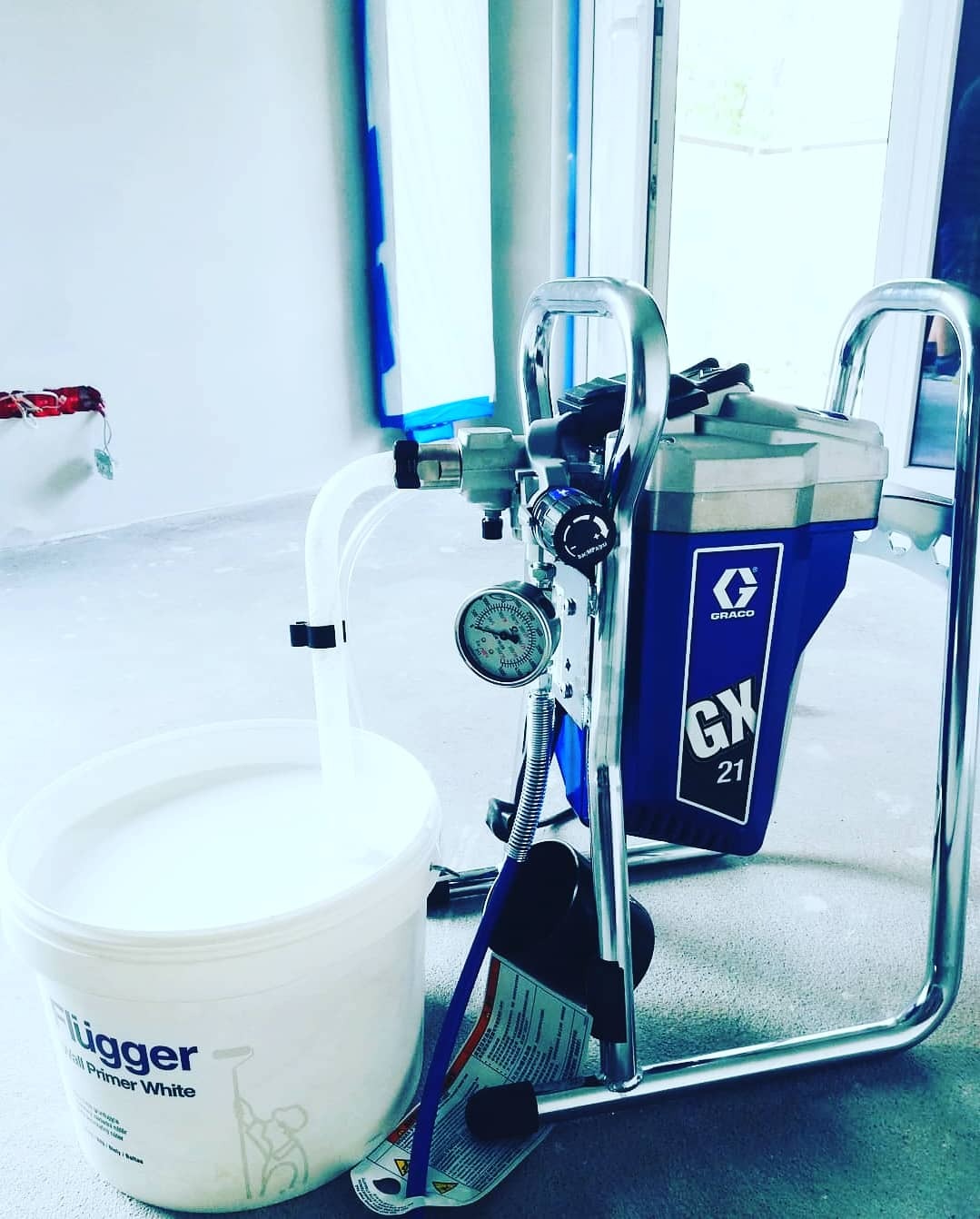 Profesjonalny agregat malarski Graco GX 21 z białym gruntem Flügger Hall Primer White w wiadrze, przygotowany do malowania ścian w pomieszczeniu zabezpieczonym taśmą malarską.