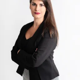 Kobieta z długimi brązowymi włosami, ubrana w czarną marynarkę i spódnicę w pepitkę, stoi na białym tle z założonymi rękami.