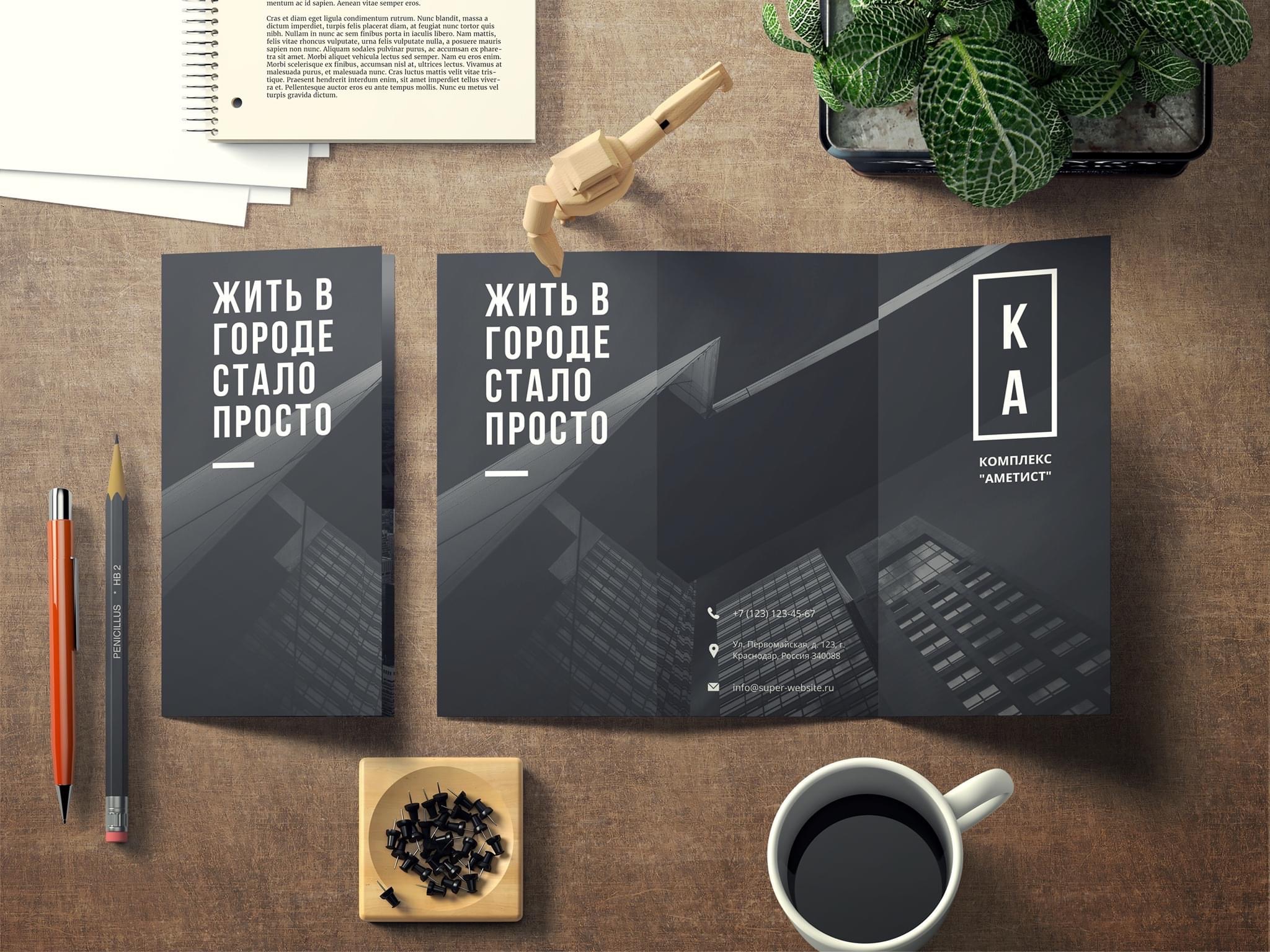 Rozłożona, grafitowa broszura trójdzielna z widokiem nowoczesnego budynku, leżąca na drewnianym biurku obok ołówków, drewnianej figurki, filiżanki kawy i notatnika.