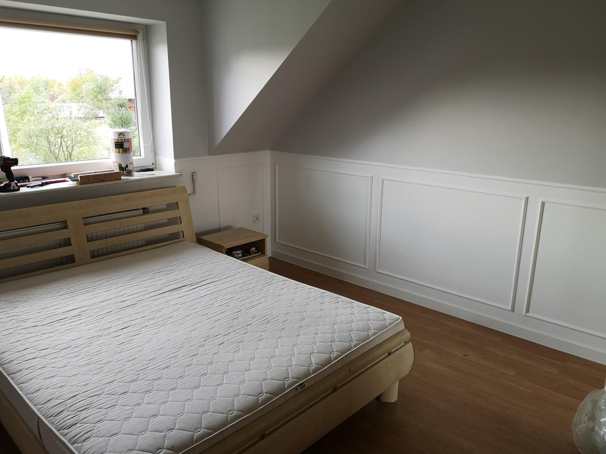 Pokój na poddaszu z łóżkiem i nową sztukaterią ścienną, widoczne narzędzia i puszki z farbą na parapecie okiennym.