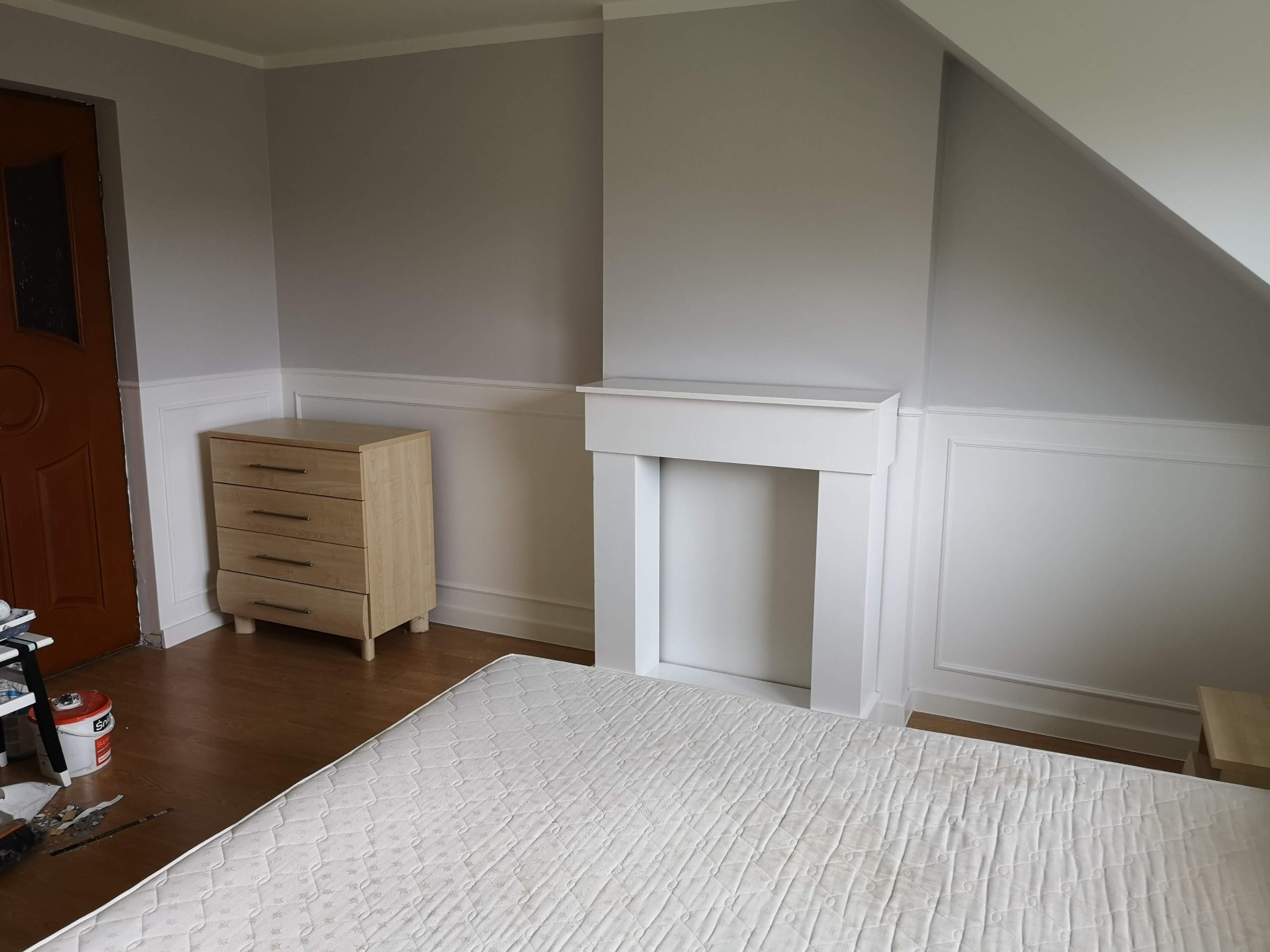 Pokój w trakcie remontu: położona podłoga z paneli, biała atrapa kominka, komoda i częściowo zakryty materac, widoczne narzędzia malarskie.