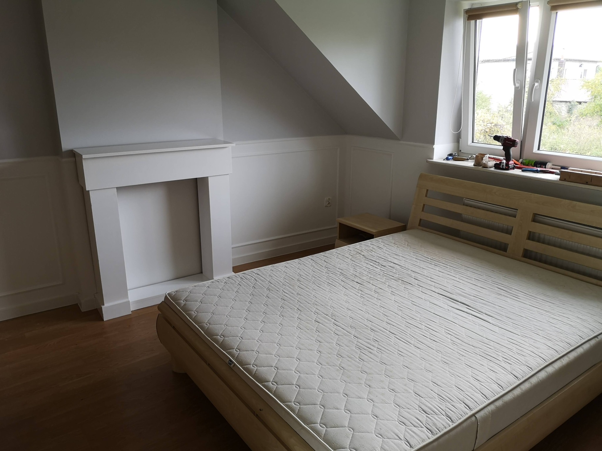 Pokój na poddaszu w trakcie remontu, z widocznym drewnianym łóżkiem bez pościeli, atrapą kominka i narzędziami leżącymi na parapecie okna.