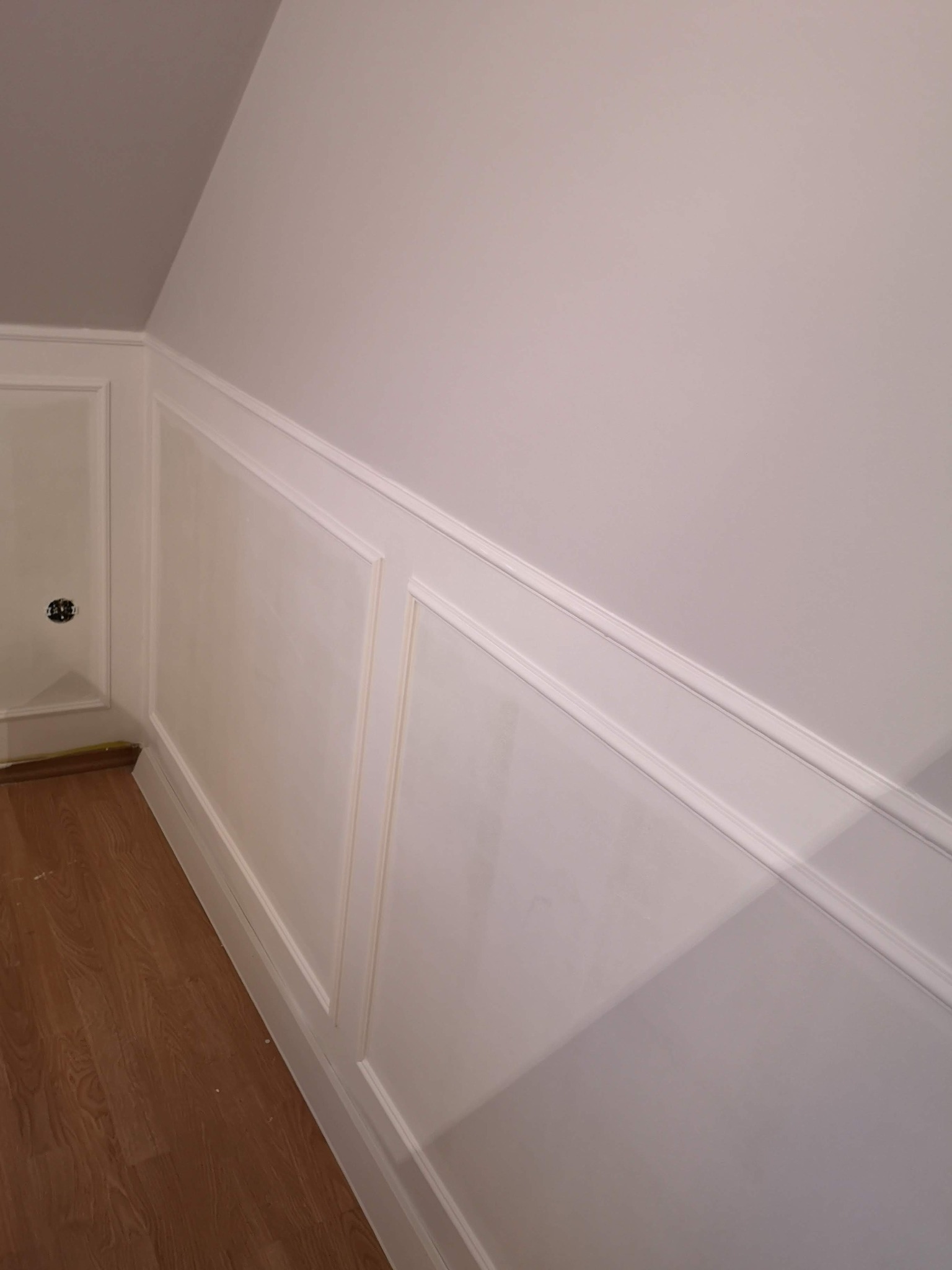 Ściana w skosie z białą sztukaterią panelową i gniazdkiem elektrycznym, widoczna podłoga z paneli drewnopodobnych.