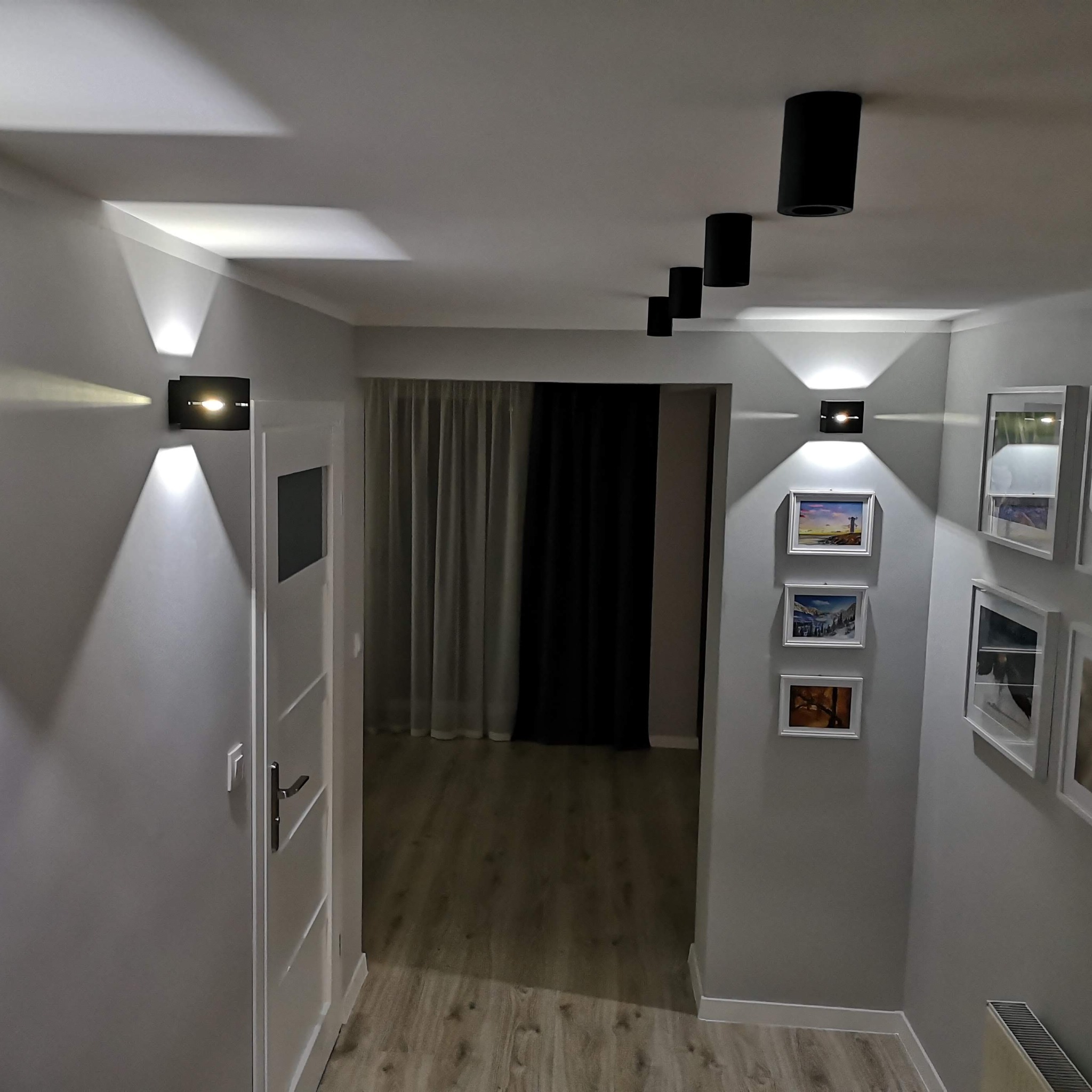Nowoczesny korytarz z szarą ścianą, drewnopodobną podłogą, białymi drzwiami i listwami przypodłogowymi, oświetlony czarnymi lampami sufitowymi i ściennymi, ozdobiony ramkami z obrazami.