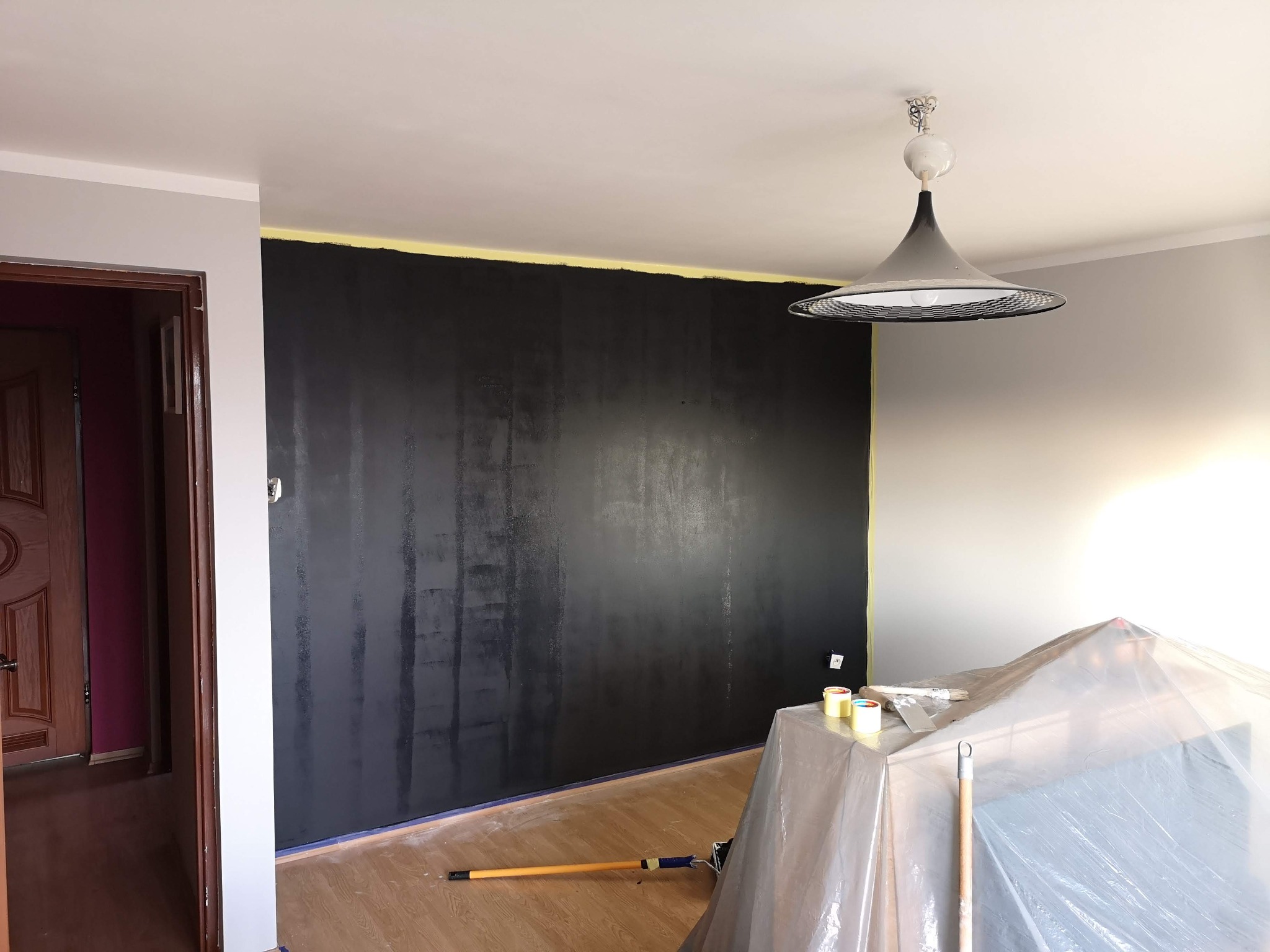 Ściana pomalowana na czarno z widocznymi smugami, zabezpieczona taśmą malarską przy suficie i podłodze, podłoga drewniana, widoczny wałek i pędzle malarskie, meble przykryte folią ochronną, lampa...