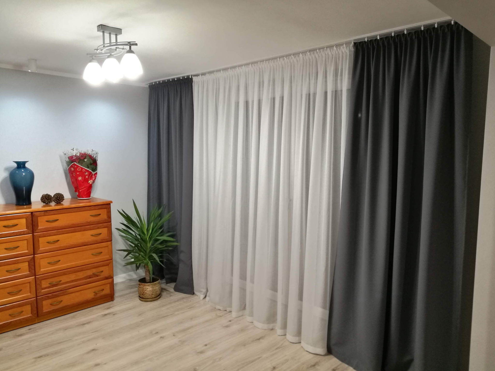 Pokój z drewnianą komodą, kwiatami, rośliną doniczkową i zasłonami: szarymi i białymi, zamontowanymi na karniszu.