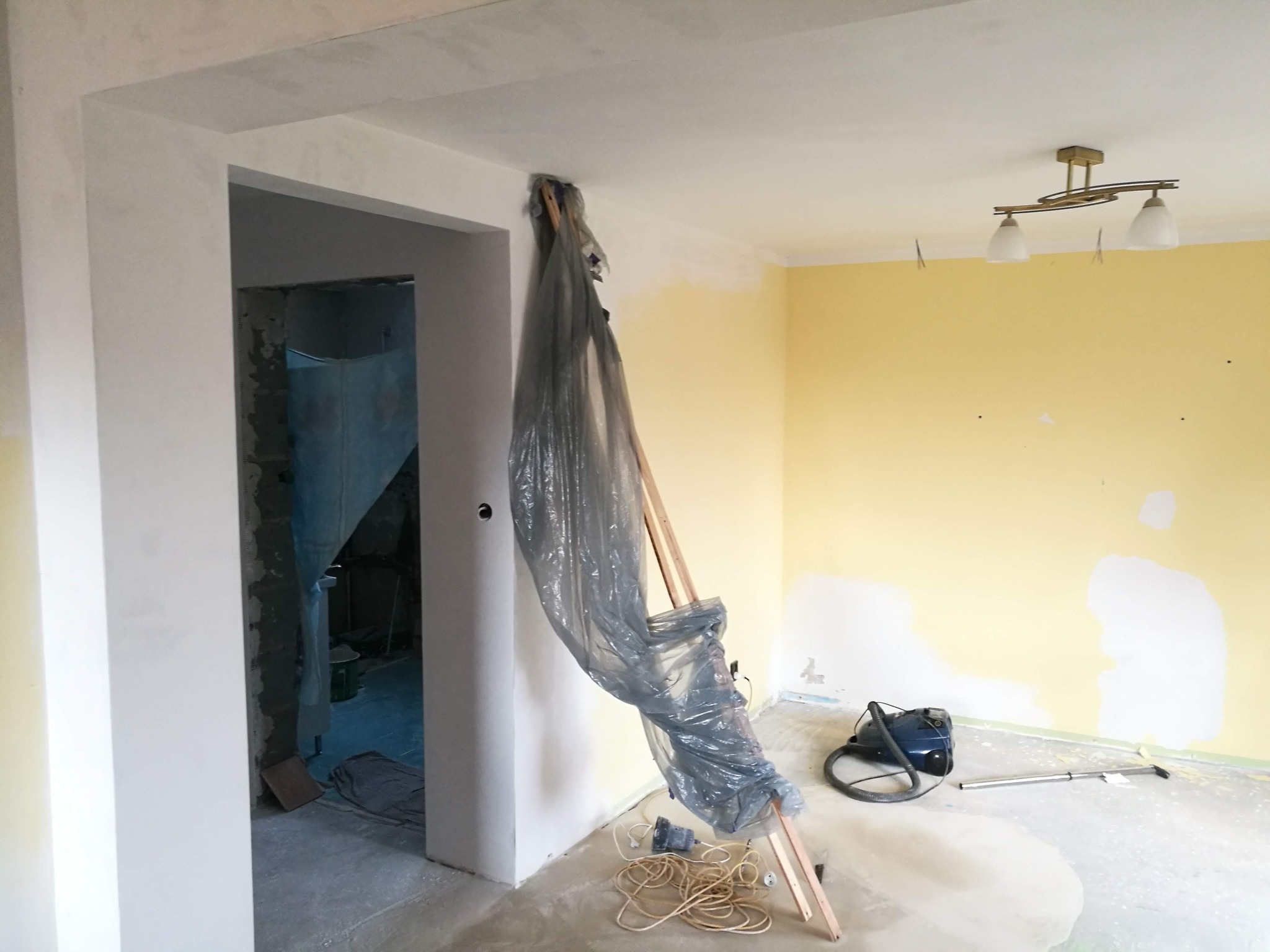 Wnętrze w trakcie remontu: żółta ściana z fragmentami świeżo nałożonej gładzi, zakryta folią drabina, odkurzacz i wisząca lampa w tle.