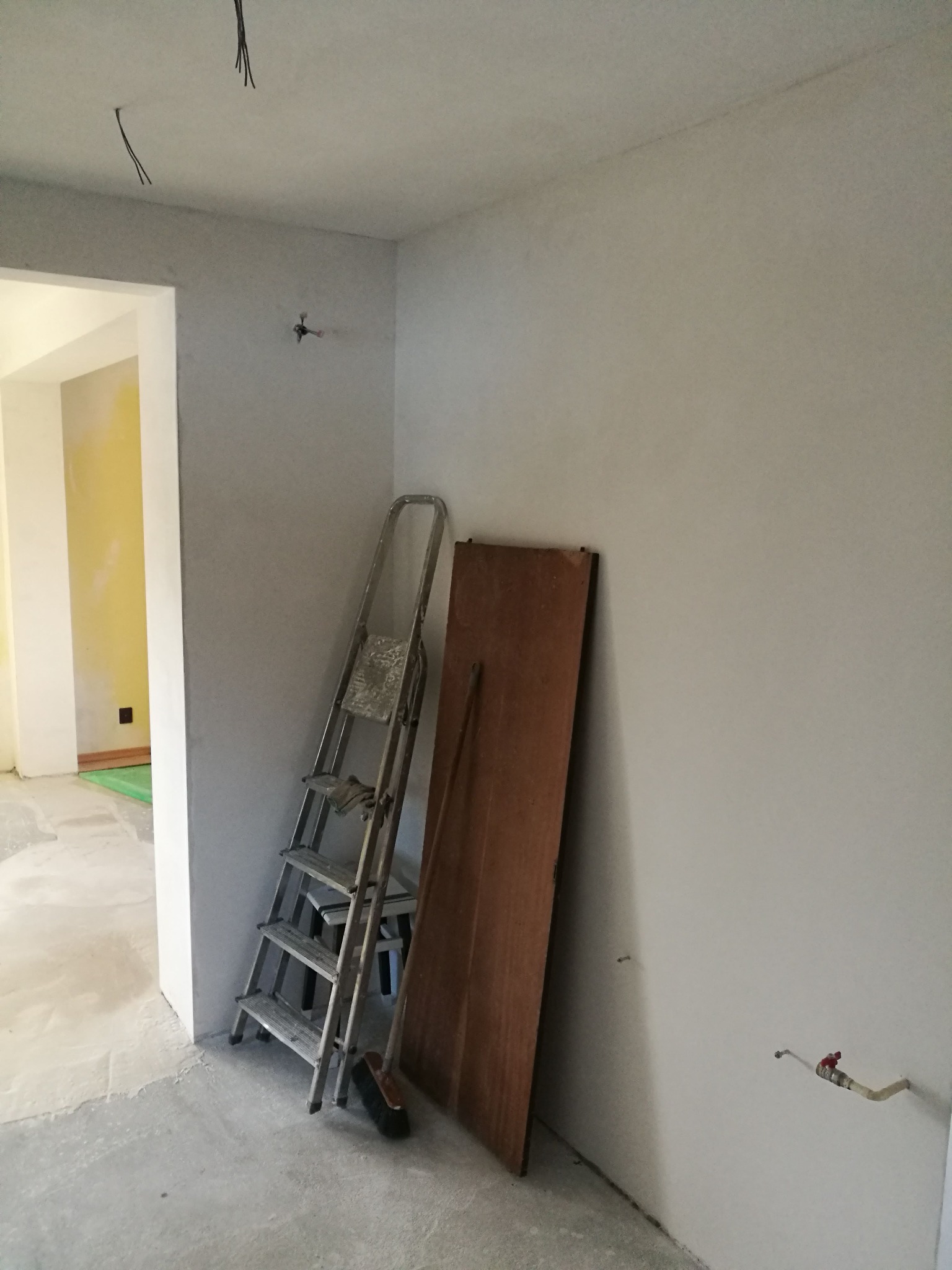 Wnętrze w trakcie remontu: surowe ściany, wystające kable z sufitu, drabina aluminiowa oparta o ścianę, drewniana płyta i szczotka leżące obok, widoczne instalacje wodne.