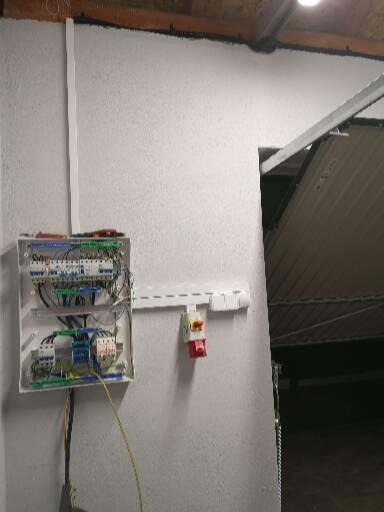 Rozdzielnica elektryczna natynkowa z widocznymi przewodami i zabezpieczeniami, zamontowana na teksturowanej ścianie obok wyłącznika nadprądowego w obudowie z czerwonym przyciskiem.