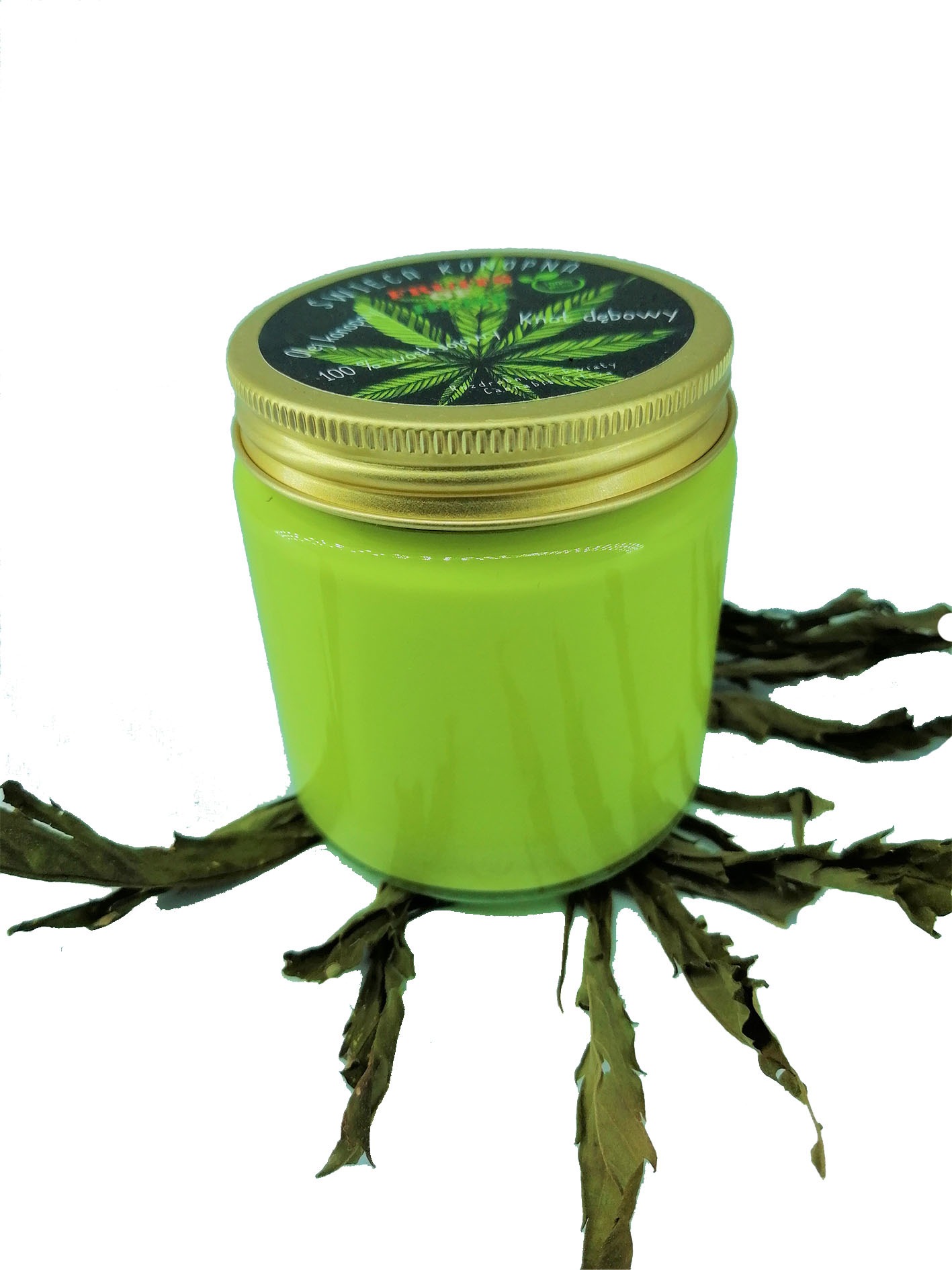Świeczka konopna w zielonym słoiczku z etykietą i złotą nakrętką, umieszczona na suszonych liściach konopi na białym tle.