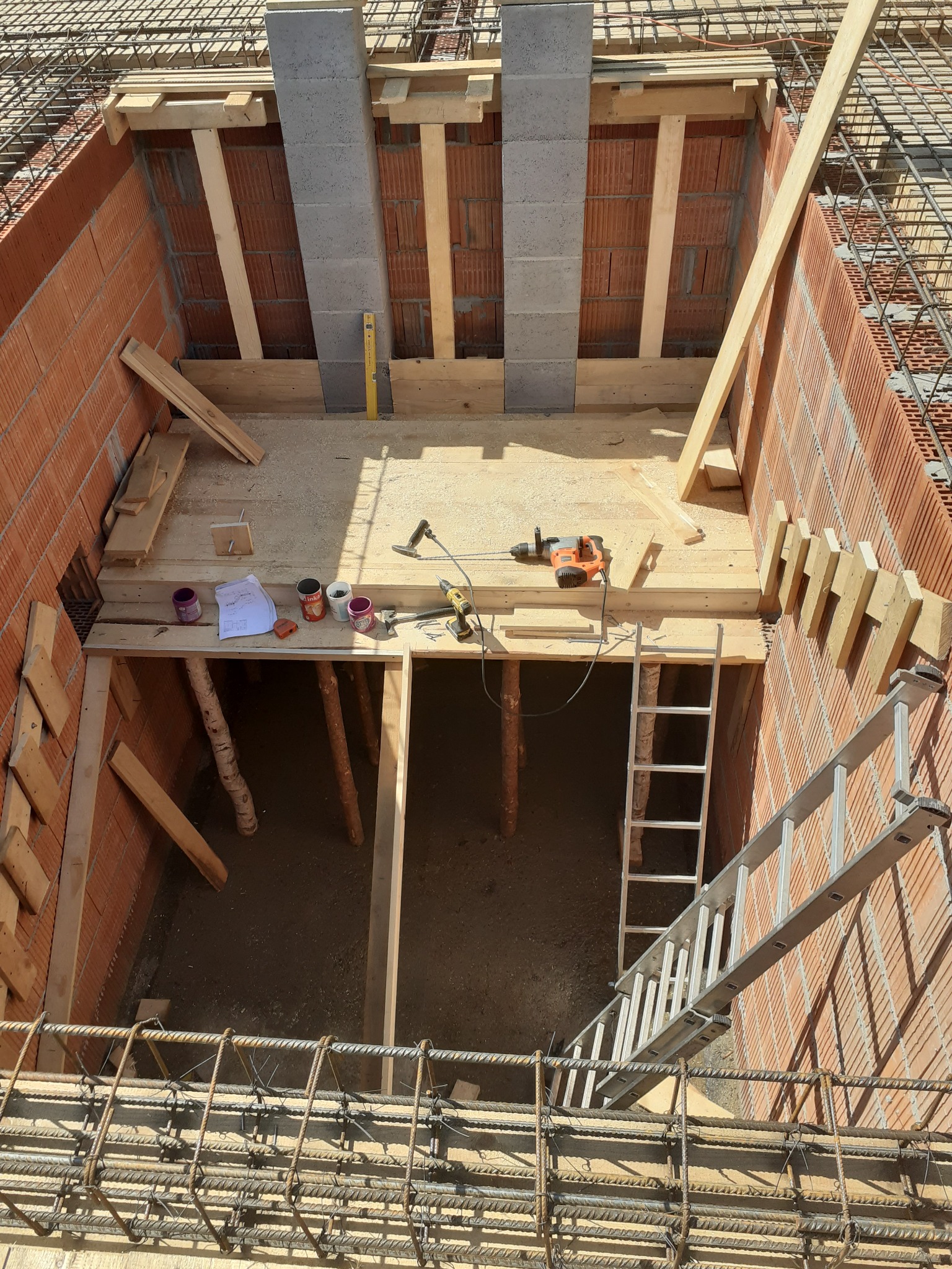 Wykop fundamentowy z deskowaniem i zbrojeniem, widoczna drabina, narzędzia i puszki z farbą na drewnianej platformie wewnątrz wykopu.