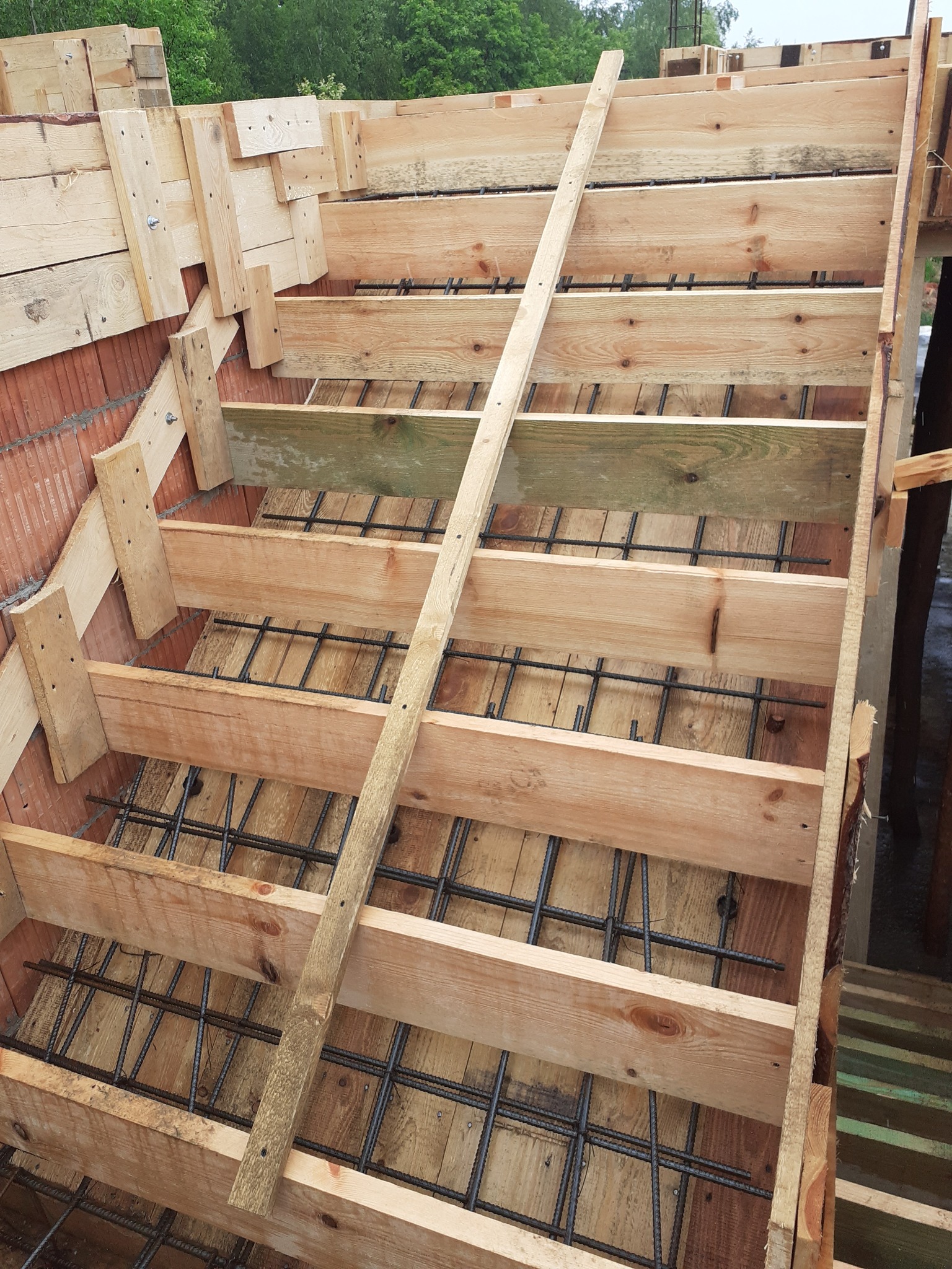 Szczegółowe ujęcie szalunku schodów z drewnianych desek i zbrojenia stalowego, widoczne połączenia śrubowe i fragment muru z cegły.