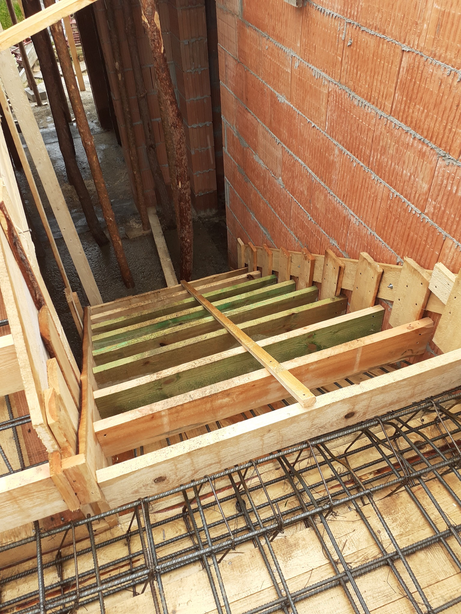 Szalunek drewniany przygotowany pod wylanie betonowych schodów, widoczna konstrukcja z belek i desek, oraz zbrojenie z prętów stalowych, ściana z czerwonej cegły dziurawki w tle.