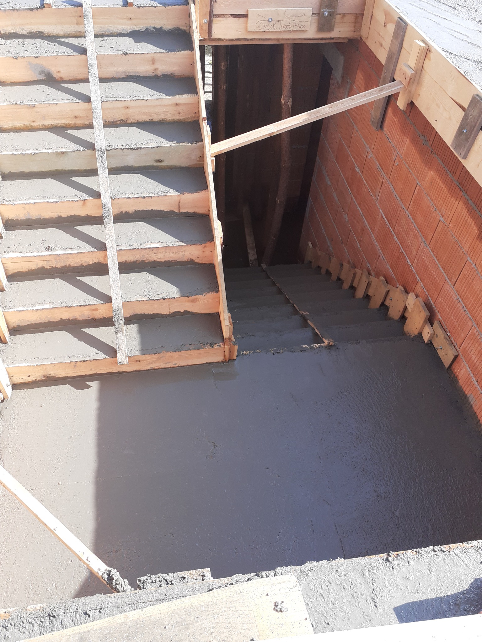 Wylewanie betonowych schodów do piwnicy z drewnianym szalunkiem i ceglaną ścianą, widoczne niedoskonałości betonu.