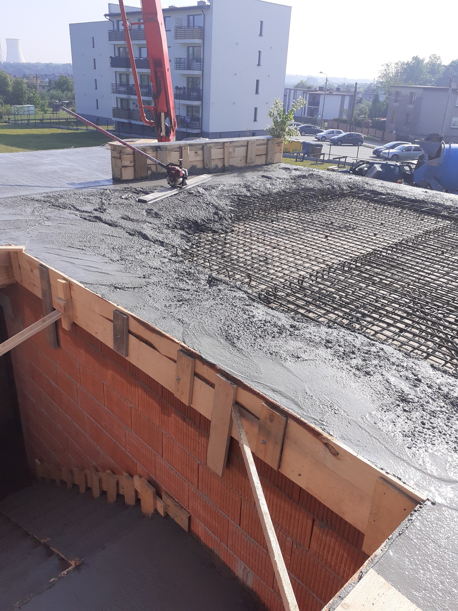 Wylewka betonu na zbrojeniu stropu budynku wielorodzinnego, z użyciem pompy do betonu i listwy wibracyjnej, widoczne szalunki i ceglana ściana.