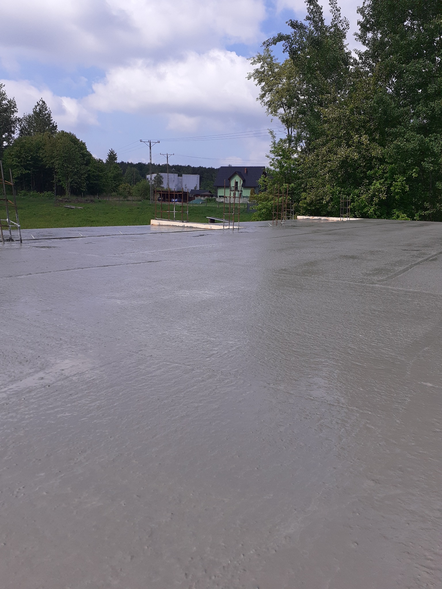 Świeżo wylana, gładka płyta betonowa z wystającymi prętami zbrojeniowymi, widok z góry na tle zieleni i zabudowań wiejskich w słoneczny dzień.