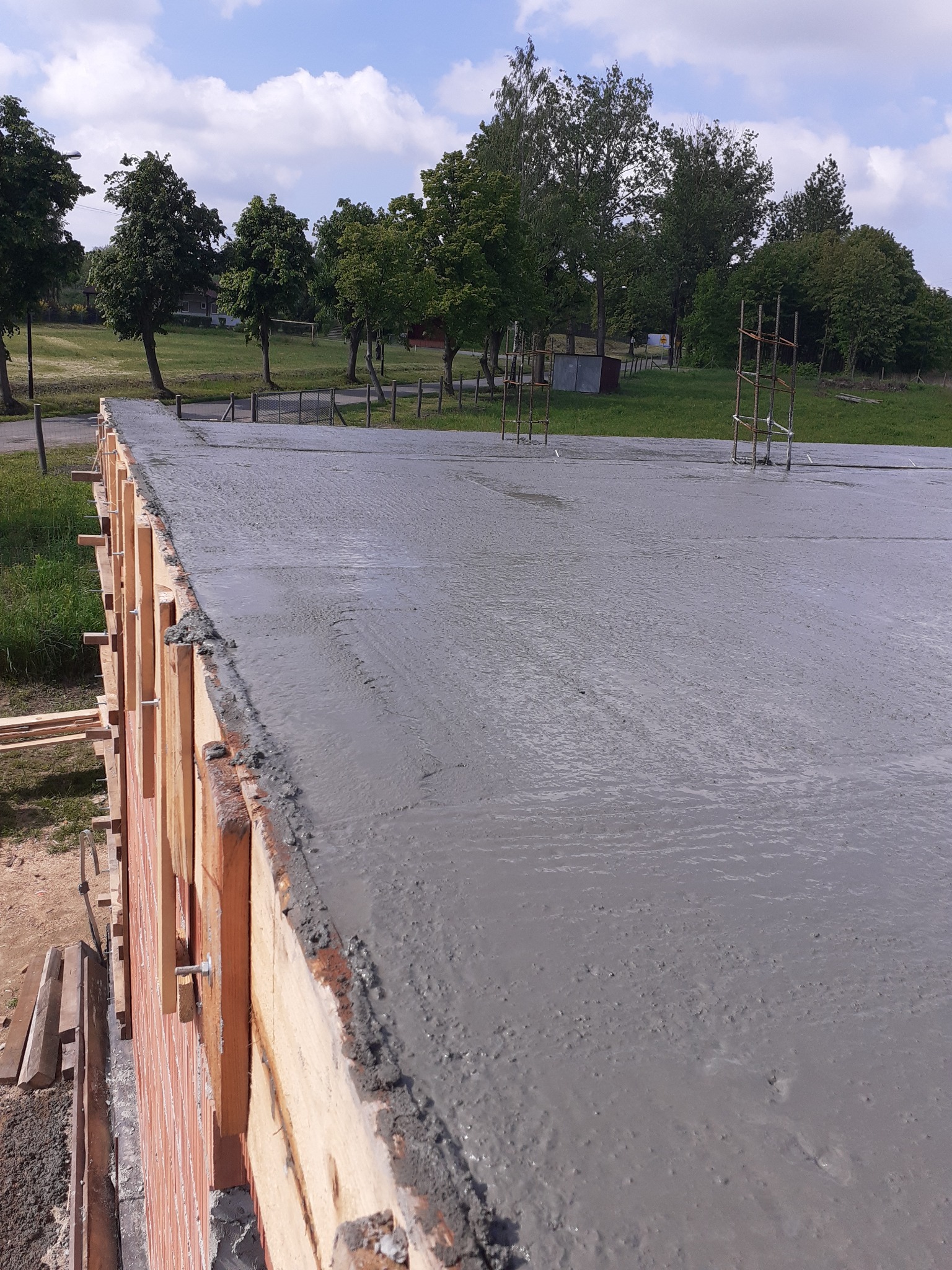 Wylany, świeży beton na przygotowanym fundamencie z widocznym szalunkiem drewnianym i wystającymi prętami zbrojeniowymi, na tle zieleni i błękitnego nieba.