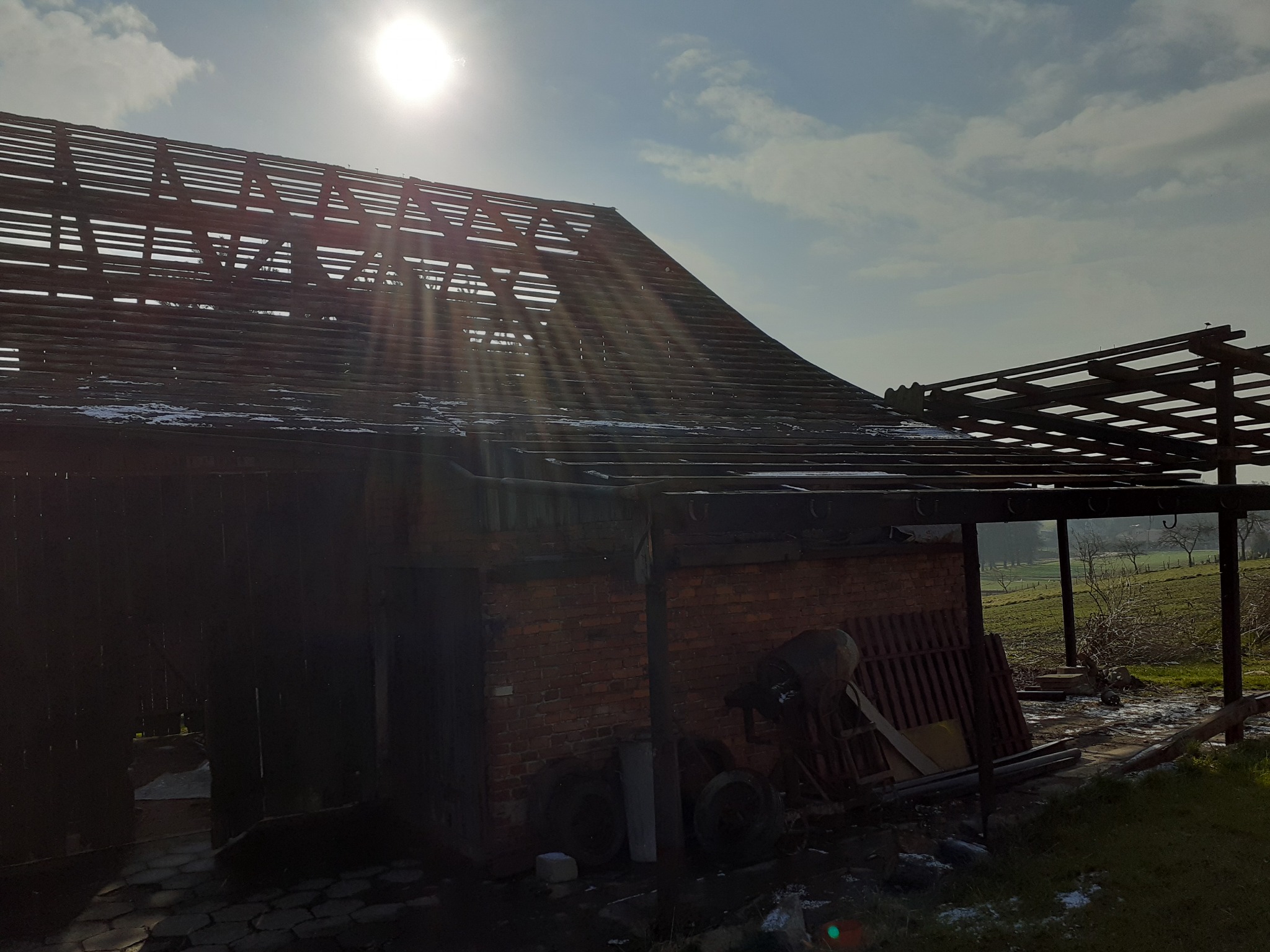 Uszkodzony, częściowo rozebrany dach starego ceglanego budynku gospodarczego w słoneczny dzień, z widocznymi elementami konstrukcji i sprzętem budowlanym w tle.