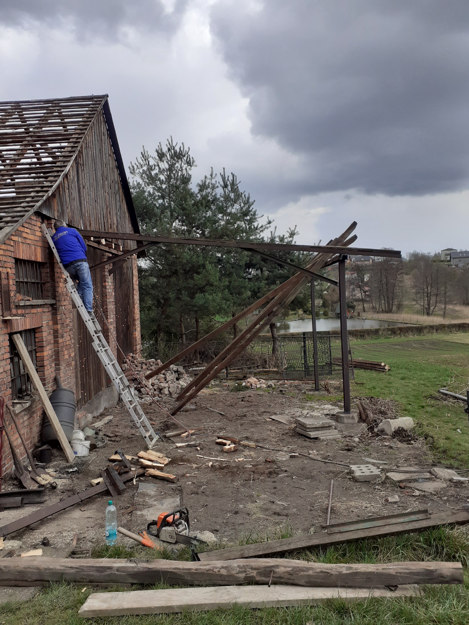 Demontaż dachu starej stodoły z cegły, pracownik na drabinie w niebieskiej bluzie, widoczne elementy konstrukcyjne, piła łańcuchowa i butelka wody na ziemi.